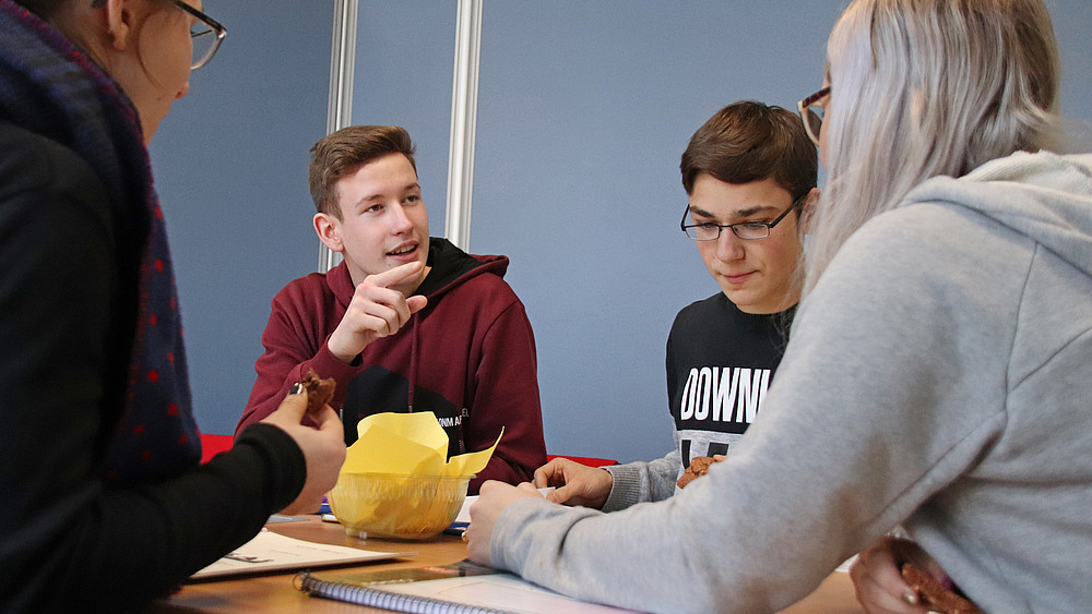 Two students are sitting around a table. There are documents and writing materials on the table. The two boys can be seen frontally, the one on the left is talking and gesticulating.