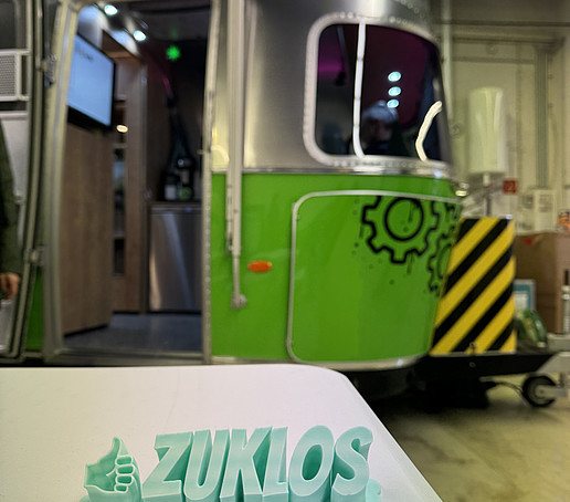 A 3D printout of the lettering ZUKLOS lies on a table in front of the AirStream trailer.