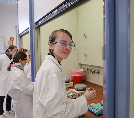 A student wearing protective goggles and a white coat stands at the extractor fan, holding a small bowl in her hand.