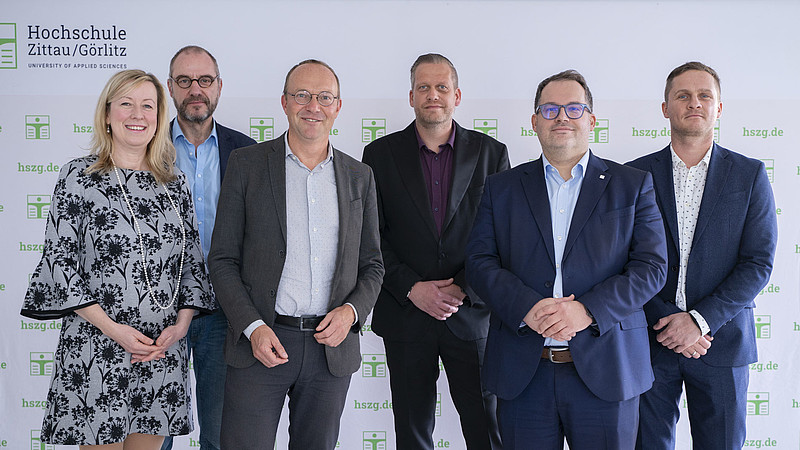 The university management, the Energy Minister and the project partners stand in front of the HSCG press wall and smile into the camera.