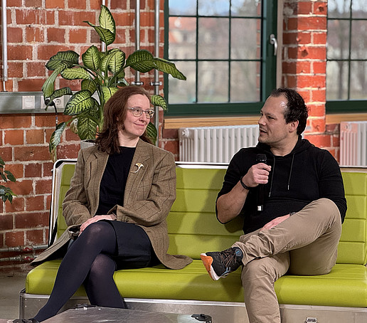 Two participants are sitting on a green sofa. The one on the right speaks into a hand-held microphone.