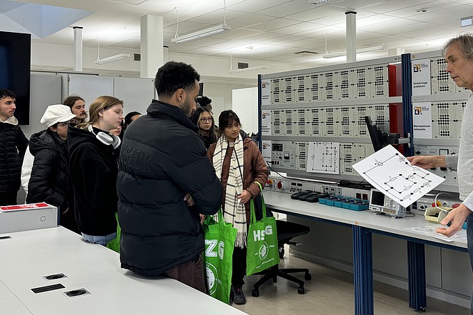 Dr. Wolfgang Menzel explains various aspects of the technical equipment to the visitors in a spacious laboratory of the Faculty of Electrical Engineering and Computer Science at the HSZG. The laboratory is modernly equipped, with numerous technical boards and devices, while the group listens attentively.