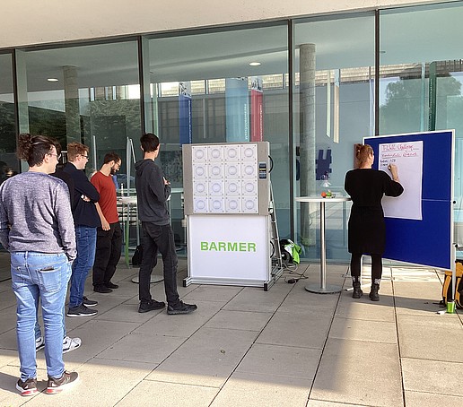 Auf dem Zittauer Campus vor Haus Z IV steht ein Barmer-Aufsteller. Eine Mitarbeiterin heftet ein Poster an eine blaue Pinwand.