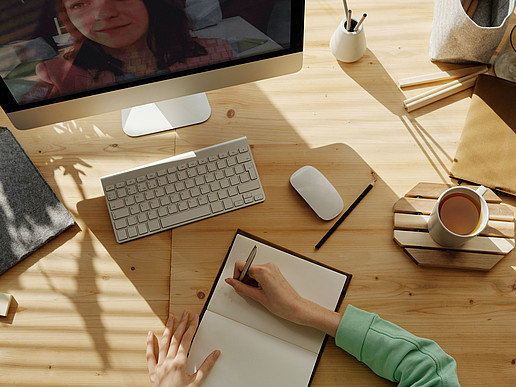 Tisch mit PC, Maus, Tastatur, Kaffeetasse, Stiften und Person die im Notizbuch schreibt. Nebenbei läuft Videokonferenz auf dem PC.