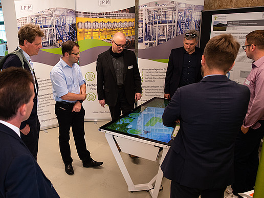 Entrepreneurs and researchers stand around a digital table and watch a presentation running there. Around them are roll-ups of the university's research work
