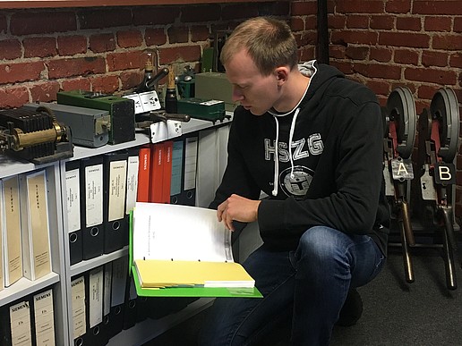 A man squats in a room in front of a shelf with many folders and holds a folder in his hand in which he reads something