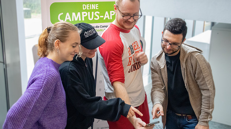 Studierende schauen gemeinsam auf ein Smartphone, im Hintergrund steht ein Aufsteller mit der Beschriftung "Deine Campus-App".
