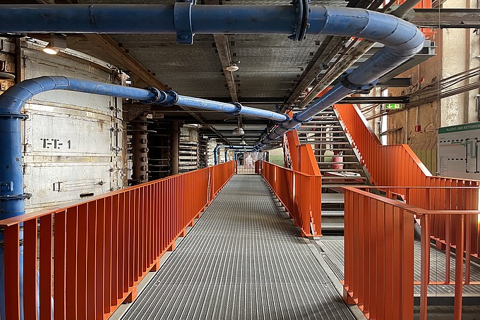 A corridor in an energy factory building with orange railings and thick blue pipes running along the ceiling.