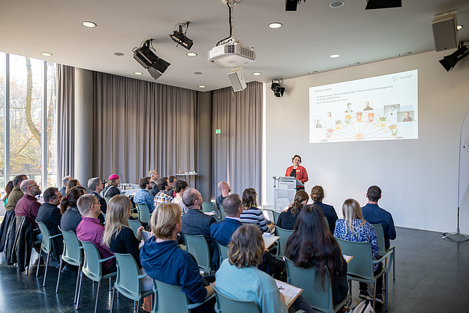 Menschen sitzen in Stuhlreihen in einer Aula und hören einer Rednerin zu.