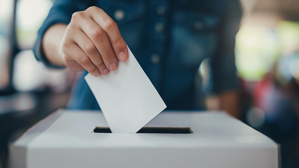 In eine weiße Wahlurne steckt eine Männerhand einen weißen Zettel.