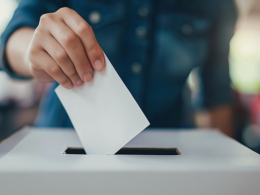 In eine weiße Wahlurne steckt eine Männerhand einen weißen Zettel.