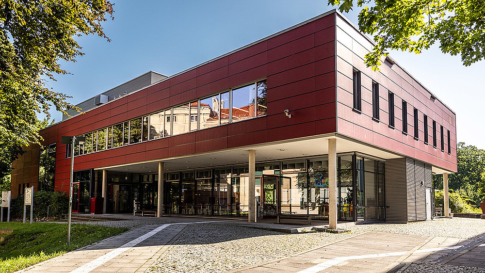 A red building on a square surrounded by trees. In the foreground, the first floor is supported by several columns.