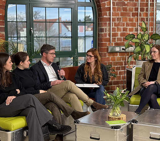 In a studio with brick walls and large windows in the background, participants sit on green sofas and chat.