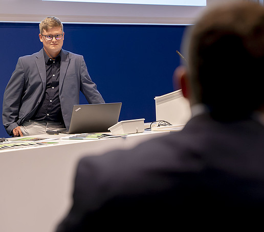 Jonas Pfeiffer steht vor einem Laptop und schaut ins Publikum. 