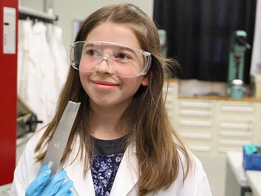 A fifth-grader wears a lab coat and safety goggles and holds a metal workpiece in her hand with a smile.