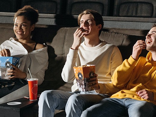 Three young moviegoers sit in the cinema eating popcorn and laughing.