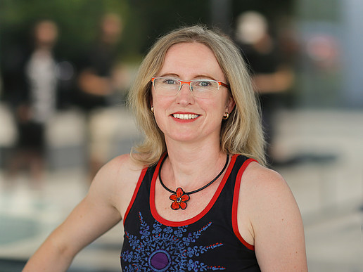 Dr. rer. nat. Kateřina Barková, eine blonde Frau mit Brille im sommerlichen Top mit roter Blumenhalskette, lächelt in die Kamera.
