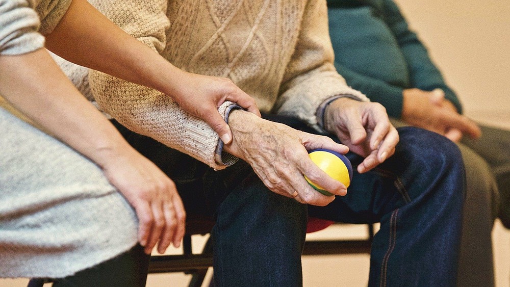 An older gentleman is sitting on a chair with a yellow ball in his hand. A younger woman is sitting next to him. She is holding and supporting his arm.