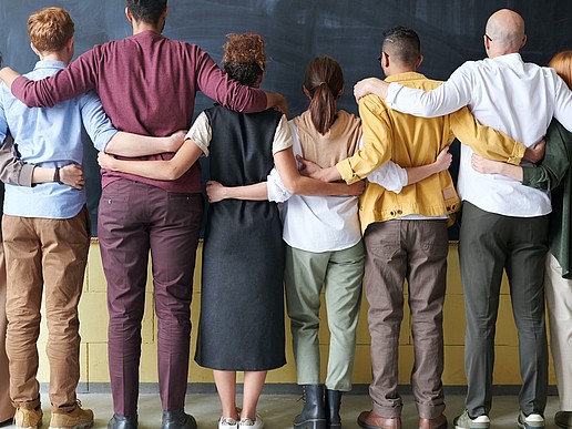 A number of young people of different ethnic backgrounds stand in a row, their backs turned to the camera, facing a blackboard. They place their hands and arms on the person standing next to them.