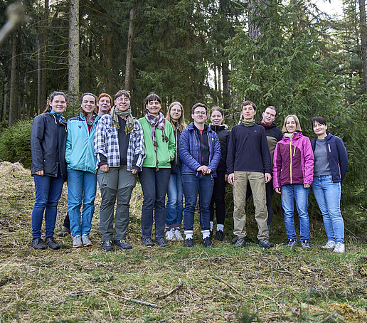 The students stand in a row on the forest floor and smile at the camera.