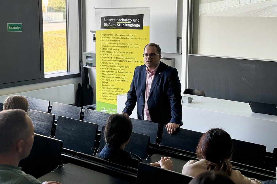 The Rector of the HSZG, Prof. Dr.-Ing. Alexander Kratzsch, stands in front of a group of prospective students from the Rahn Studienkolleg Leipzig and welcomes them to the lecture hall. In the background you can see a banner with courses on offer and a presentation screen.