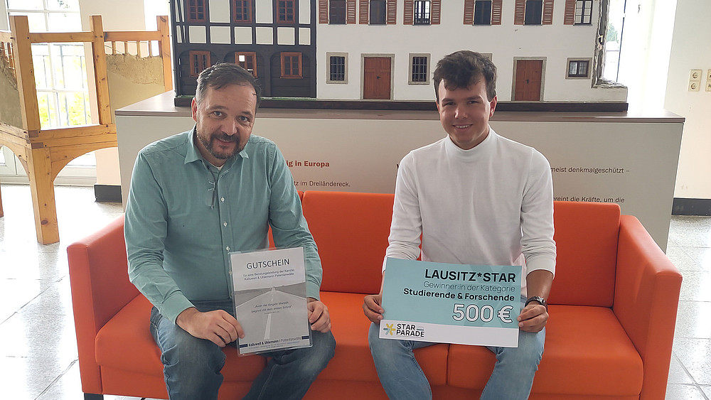 David Sauer and a student sit on a couch and hold a certificate and a check in front of them. In the background is a model of a half-timbered house.