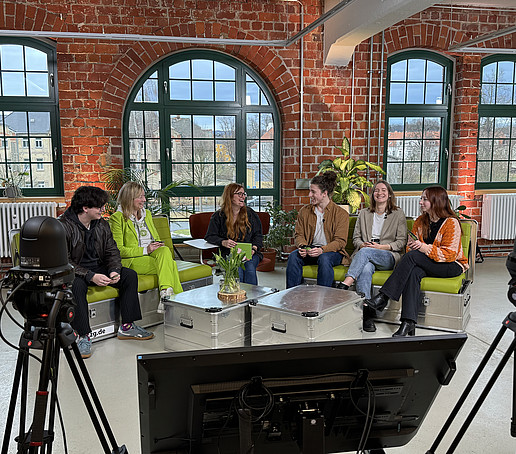 In a studio with brick walls and large windows in the background, participants sit on green sofas and chat.