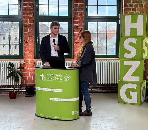 A contributor and the presenter stand behind an HSZG counter in the studio. On the left in the foreground is a black camera. Four HSZG cubes are piled up to the right of the counter.