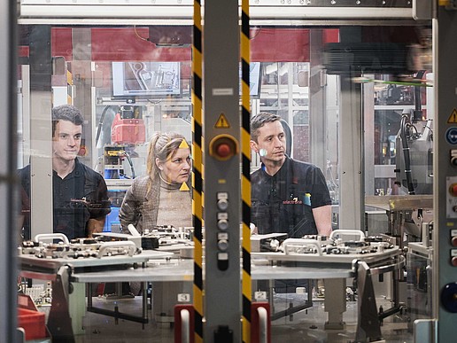 Three people observe a production plant behind a pane of glass.