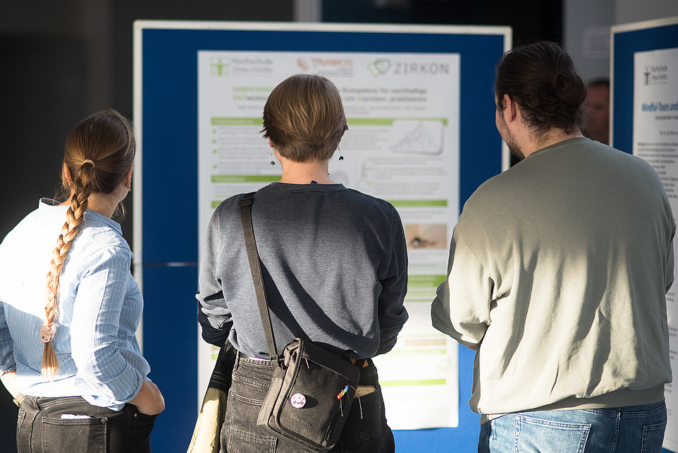 Three people stand in front of a poster wall and look at a poster with research results on it.