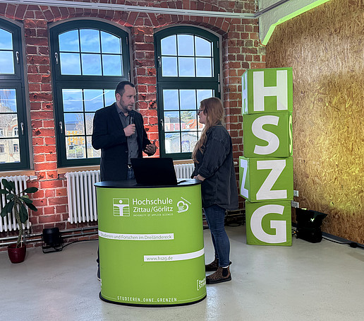 A contributor and the presenter stand behind an HSZG counter in the studio. On the left in the foreground is a black camera. Four HSZG cubes are piled up to the right of the counter.