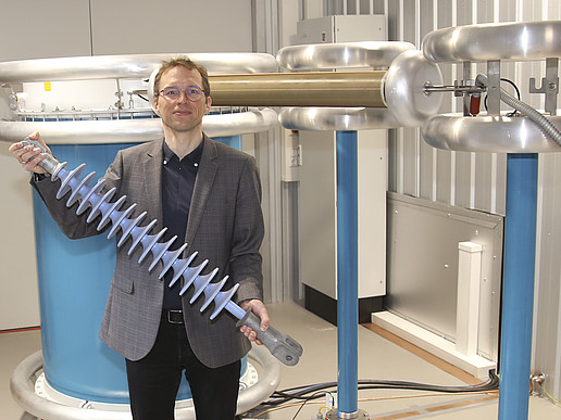 A professor stands in front of a voltage system and holds a component in his hand.
