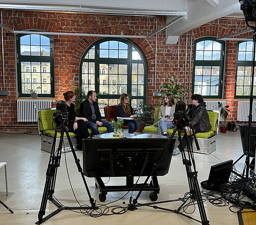 In a studio with brick walls and large windows in the background, participants sit on green sofas and chat.