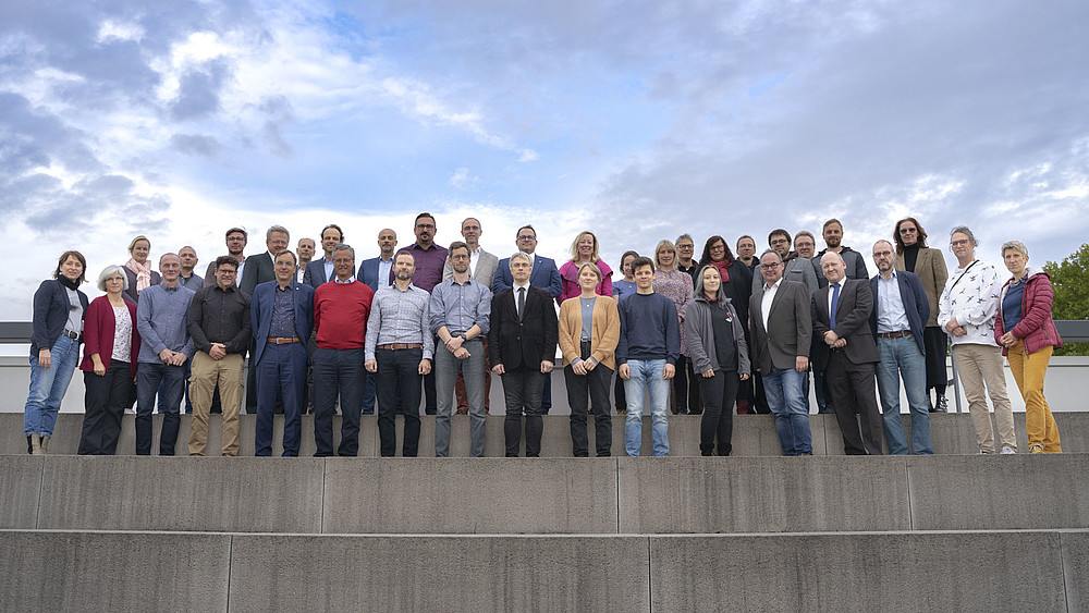 On a roof terrace, the members of the Extended Senate stand in several rows on stone steps.