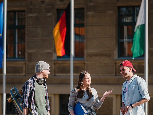 The European, German and Saxon flags fly in front of a university building in Görlitz. Three students are talking in front of it.