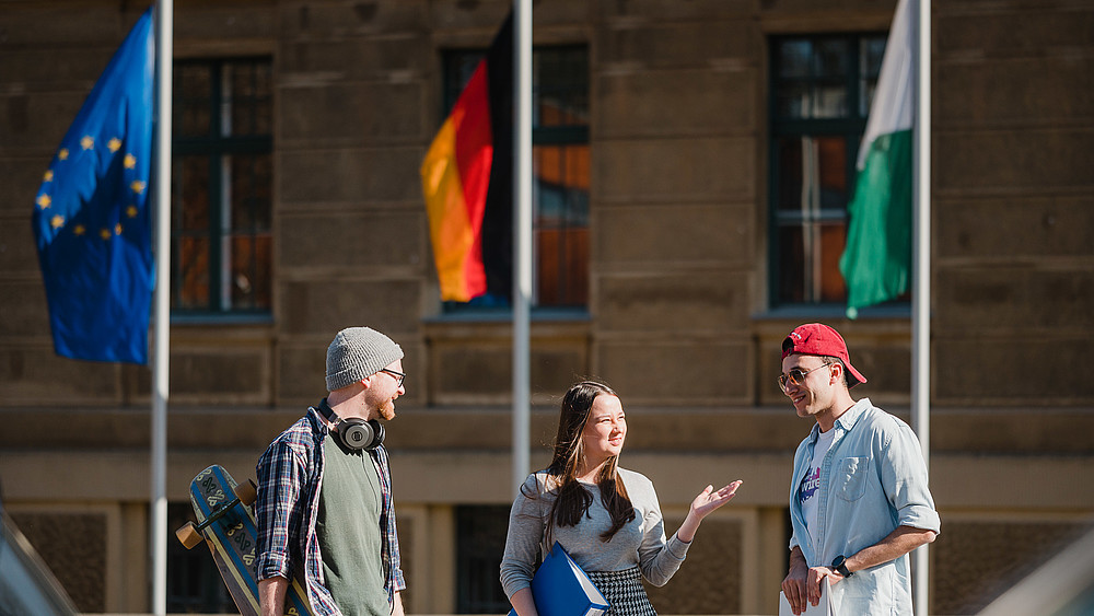 The European, German and Saxon flags fly in front of a university building in Görlitz. Three students are talking in front of it.