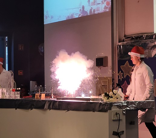 Two lab technicians in Christmas hats experiment on stage. Fireworks light up in the middle of the table.