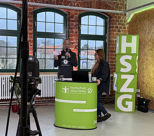 A contributor and the presenter stand behind an HSZG counter in the studio. On the left in the foreground is a black camera. Four HSZG cubes are piled up to the right of the counter.