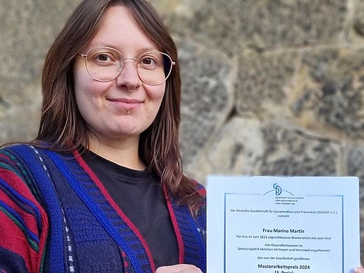 A student with glasses stands in front of a stone wall and holds a certificate smiling at the camera.