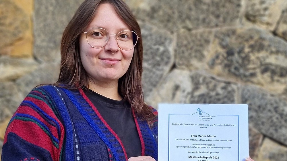 A student with glasses stands in front of a stone wall and holds a certificate smiling at the camera.