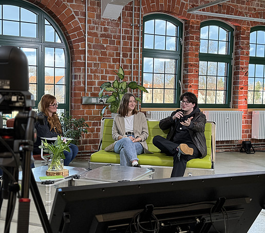 Two participants are sitting on a green sofa. The one on the right speaks into a hand-held microphone.