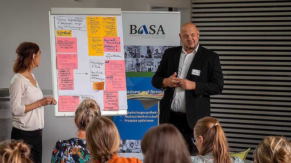 A professor stands in front of full rows of seats and explains something using a board covered with slips of paper.