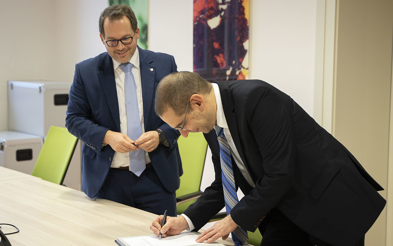 Dr. Zeller stands bent over a table and signs the honorary professorship certificate. The Rector stands behind him, smiling and watching him.