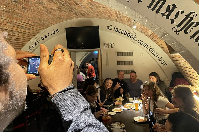 A group of college students sit at a table in the Dezi Bar and have a happy evening. One participant holds a camera to capture the memories, while other guests sit and eat together in the background.