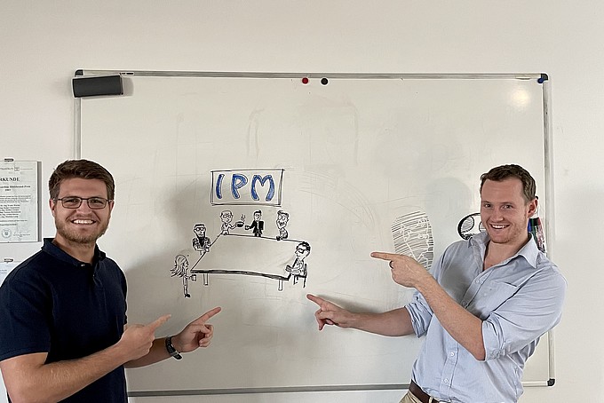 Jan-Hendrik and Karl point to the inscription "IPM" on a whiteboard.