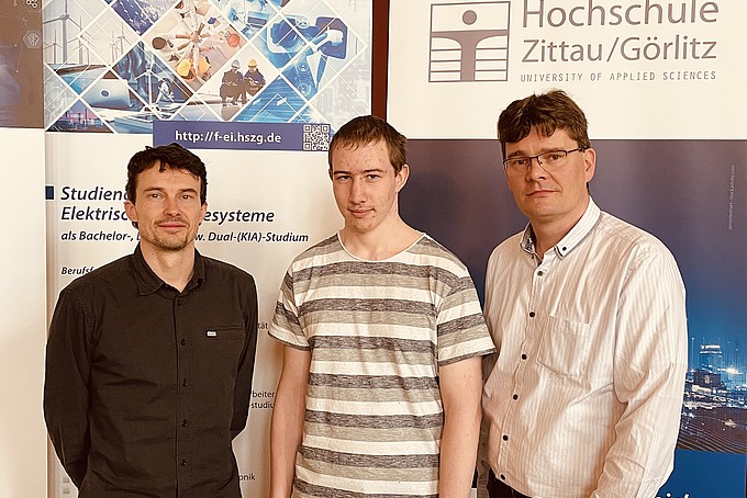 Three men stand in front of a billboard with the words "Hochschule Zittau/Görlitz" on it