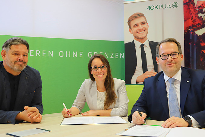 (from left to right) Mario Hanke and Linda Zölfel from AOK Plus and Rector Prof. Dr.-Ing. Alexander Kratzsch have the cooperation agreement on the table in front of them and smile at the camera.
